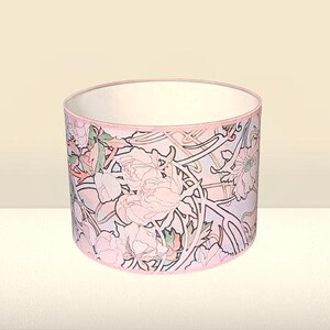 May include: A cylindrical drum lampshade featuring a repeating floral pattern with soft pink peonies, green leaves, and dark brown outlines on a light background. The top and bottom edges are finished with a thin, solid pink fabric trim.