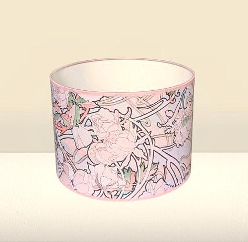 May include: A cylindrical drum lampshade featuring a repeating floral pattern with soft pink peonies, green leaves, and dark brown outlines on a light background. The top and bottom edges are finished with a thin, solid pink fabric trim.