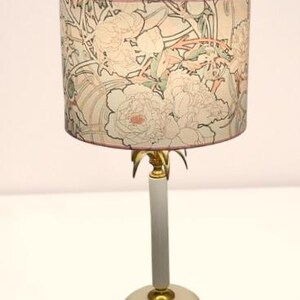 May include: A table lamp featuring a cylindrical lampshade decorated with a floral pattern in shades of pink, green, and cream. The lamp has a slender, light gray cylindrical stem, a circular base, and gold-toned metal accents at the joints.