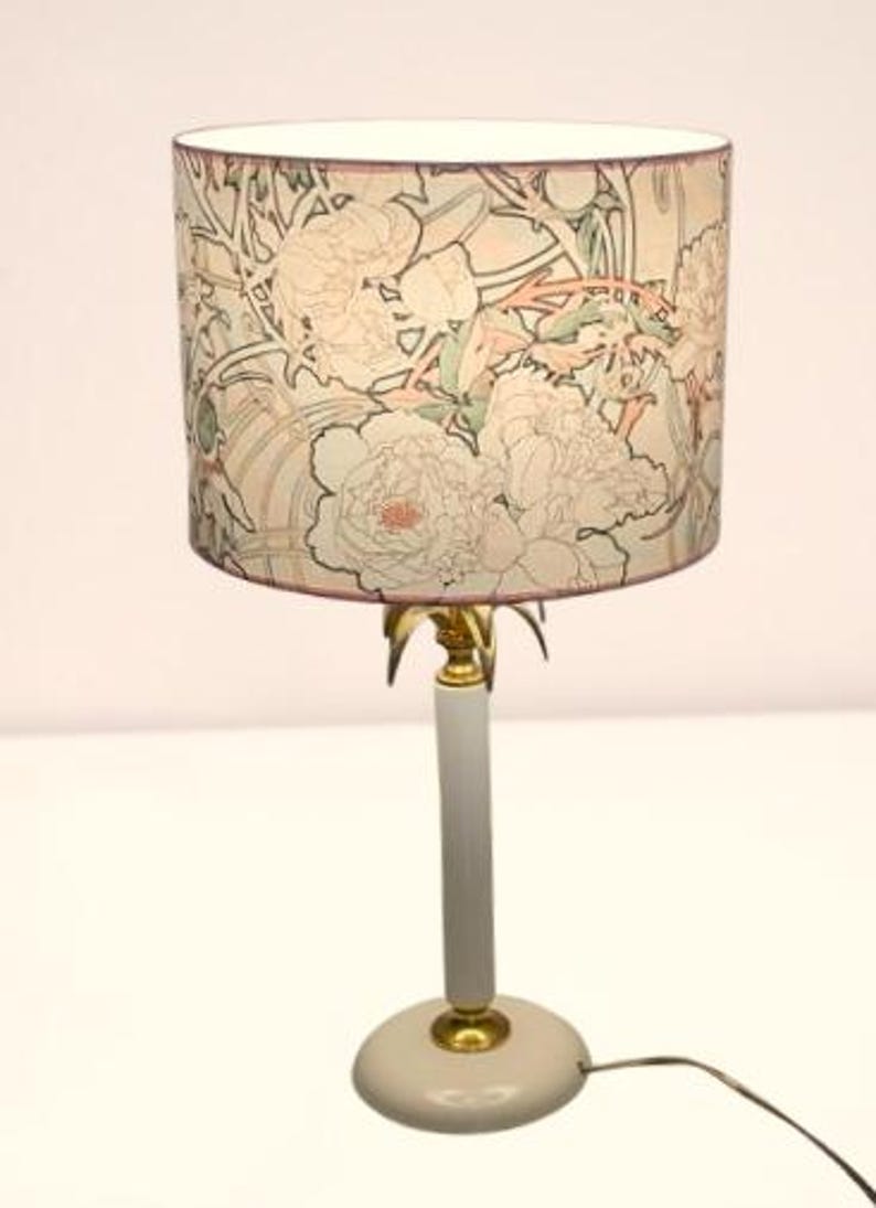 May include: A table lamp featuring a cylindrical lampshade decorated with a floral pattern in shades of pink, green, and cream. The lamp has a slender, light gray cylindrical stem, a circular base, and gold-toned metal accents at the joints.
