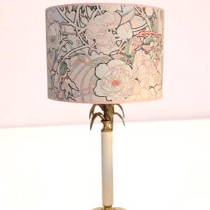 May include: A table lamp featuring a cylindrical lampshade with a light-colored background and a black-lined floral pattern in shades of pink and green. The lamp base is a slender white column with gold-toned metal accents and a round white base.