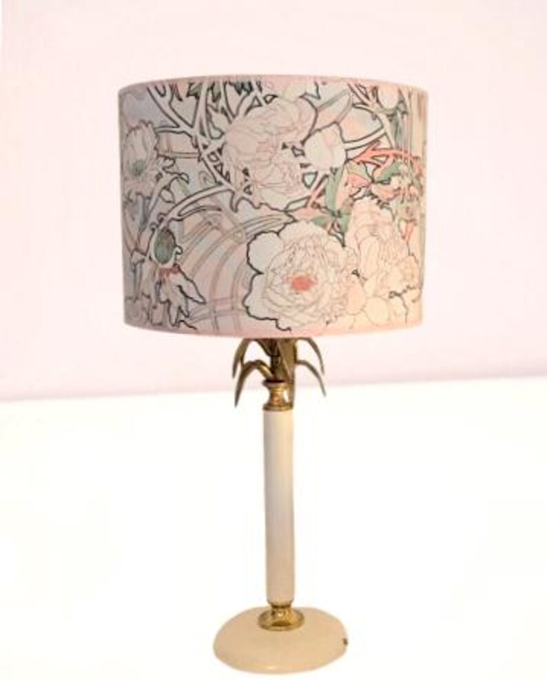 May include: A table lamp featuring a cylindrical lampshade with a light-colored background and a black-lined floral pattern in shades of pink and green. The lamp base is a slender white column with gold-toned metal accents and a round white base.