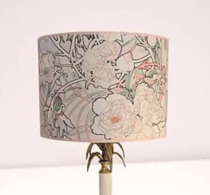 May include: A cylindrical lamp shade featuring a light-colored background with a delicate black line art pattern of blooming flowers and foliage, accented with soft pink and muted green tones, supported by a metallic gold-toned base structure.