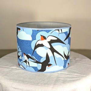 May include: Cylindrical lampshade with a blue background featuring white clouds and stylized black swallows with orange and white accents. The lampshade has a light blue trim and a silver interior.