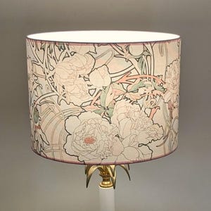 May include: A cylindrical lamp shade featuring a repeating floral pattern of large peonies and winding stems in soft shades of pink, sage green, and cream on a light background, supported by a gold-toned metal base with pointed accents.
