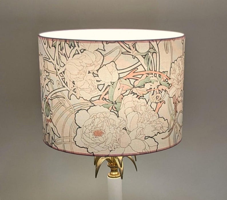 May include: A cylindrical lamp shade featuring a repeating floral pattern of large peonies and winding stems in soft shades of pink, sage green, and cream on a light background, supported by a gold-toned metal base with pointed accents.