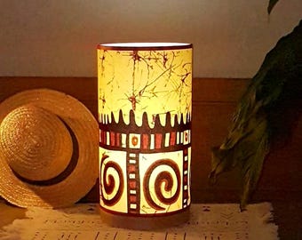 May include: A cylindrical table lamp with a warm, illuminated cream-colored shade featuring a dark brown and red geometric pattern of spirals and vertical lines, accented by a subtle, thin, web-like crackle texture across the upper section.