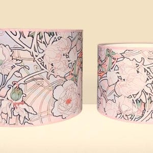 May include: Two cylindrical drum lampshades of different heights featuring a repeating floral pattern with light pink, soft green, and white outlines on a pale background. Each shade has a thin pink trim along the top and bottom edges.