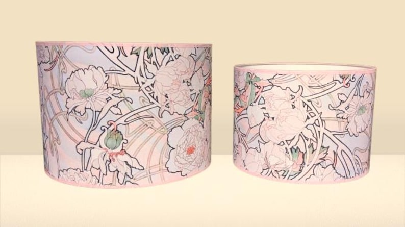 May include: Two cylindrical drum lampshades of different heights featuring a repeating floral pattern with light pink, soft green, and white outlines on a pale background. Each shade has a thin pink trim along the top and bottom edges.