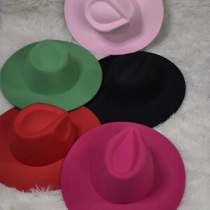 Op de afbeelding: Vijf vilt fedora hoeden in verschillende kleuren: roze, groen, zwart, rood en fuchsia. De hoeden zijn in een cirkel op een wit, pluche oppervlak gerangschikt.