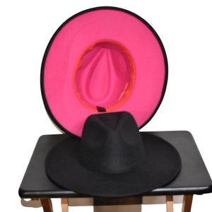 Op de afbeelding: Een zwarte fedora-hoed met een roze en oranje rand. De hoed ligt op een zwart oppervlak.