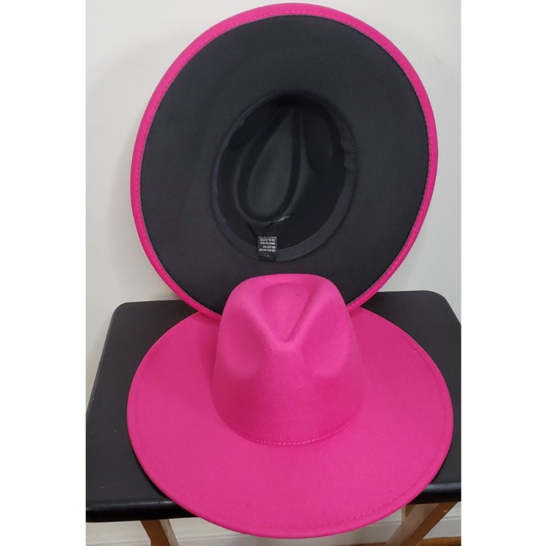 Op de afbeelding: Een roze fedora-hoed met een zwarte viltvoering. De hoed heeft een brede rand en een klassieke fedora-vorm.
