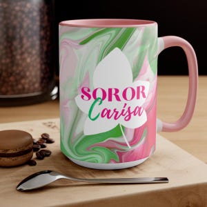 May include: A pink and green swirl design mug with a white leaf and the text "SOROR Carisa" in pink. The mug has a pink handle.