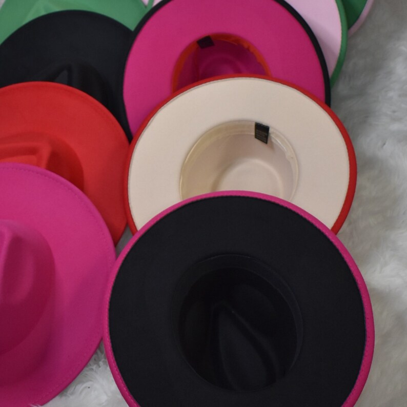 Op de afbeelding: Een stapel felgekleurde vilt fedora hoeden met verschillende gekleurde banden. De hoeden zijn in een cirkel gerangschikt met de bovenste hoed die naar de camera wijst. De hoeden zijn allemaal in verschillende tinten roze, rood, zwart en groen.