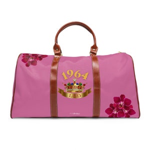 Puede incluir: Bolsa de viaje de lona rosa con detalles de cuero marrón. La bolsa presenta una corona dorada con joyas y el texto "1964 LOD". La bolsa está decorada con flores de orquídea rosa.