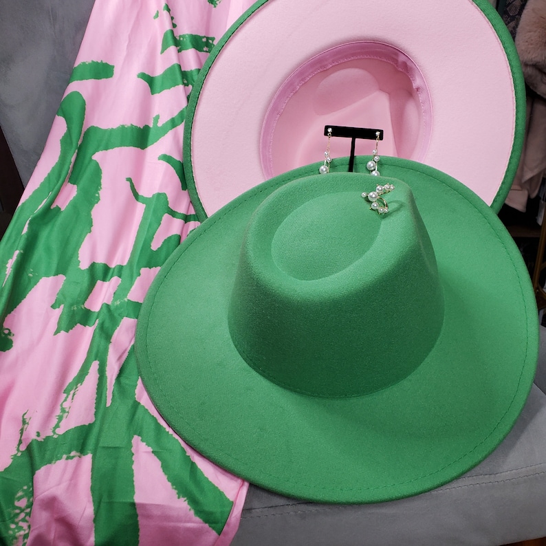 Op de afbeelding: Een groene fedora-hoed met daaronder een roze hoed. De hoeden liggen op een roze en groene stof met een patroon.