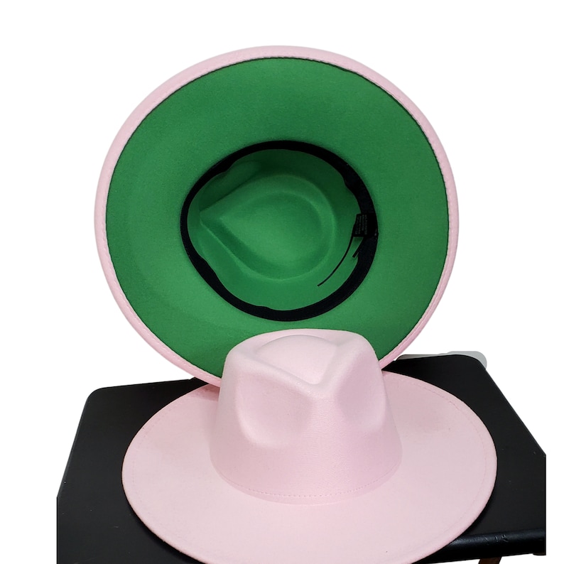 Op de afbeelding: Een roze fedora-hoed met een groene viltvoering. De hoed heeft een brede rand en een licht ingedeukte kroon.