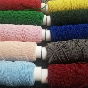 Puede incluir: Una colección de diez carretes de hilo en varios colores, incluyendo rojo, verde, rosa, beige, azul, gris y rojo oscuro. Los carretes están dispuestos en una fila, con los extremos del hilo visibles.