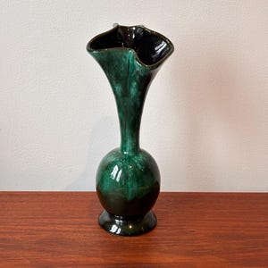 May include: A tall, green ceramic vase with a flared, ruffled top and a bulbous midsection. The vase has a glossy finish with dark green and black tones. It stands on a round base and is displayed on a wooden surface.