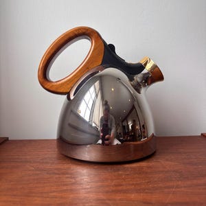 Può includere: Una teiera lucida in acciaio inossidabile con manico in legno e beccuccio dorato. La teiera riflette l'ambiente circostante. È appoggiata su una superficie di legno. La teiera è alta circa 20 cm.