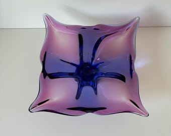 Puede incluir: Un cuenco de vidrio morado y azul con una forma orgánica única. El cuenco tiene una base ancha y plana y un centro que se eleva hasta un punto. El vidrio es iridiscente y tiene un brillo sutil.