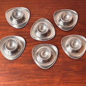 Könnte beinhalten: Sechs silberfarbene Eierbecher mit einem einzigartigen dreieckigen Design. Jeder Becher hat eine zentrale, abgerundete Halterung für das Ei, die sich in einer flachen, ausgestellten Basis befindet. Die Becher sind auf einer dunklen Holzoberfläche angeordnet.