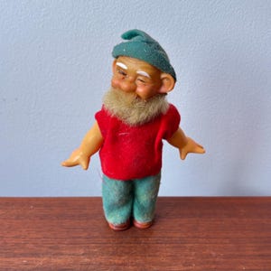 May include: A vintage gnome figurine with a green hat, red shirt, and green pants. The gnome has a white beard and is smiling. The figurine is standing on a wooden surface.
