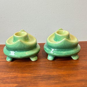 May include: A pair of green ceramic candle holders. Each has a round base with three small feet and a central cup for a candle. The candle holders have a glossy finish and an organic shape. The background is a neutral colour.
