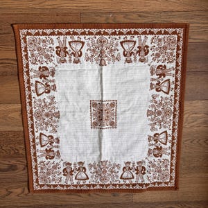 May include: A white tablecloth with a brown border featuring a repeating pattern of stylized figures and floral motifs. The border is trimmed with a brown fringe.