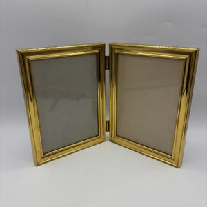 May include: A gold-toned double picture frame with two openings. The frame is hinged in the middle and can be opened to reveal the two openings.