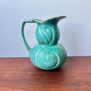 May include: A small, turquoise-colored ceramic pitcher with a bulbous base and a floral-inspired design. The pitcher has a curved handle and a flared spout. The surface has a textured, mottled appearance, and it sits on a wooden surface.