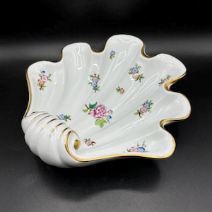 May include: A white porcelain seashell-shaped dish with a gold rim and a floral pattern in shades of pink, blue, and green.