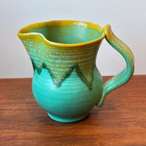 May include: A ceramic pitcher featuring a rounded body and a curved handle. The exterior is finished with a vibrant turquoise glaze on the lower portion, transitioning to a textured, mottled mustard yellow glaze along the rim and handle.