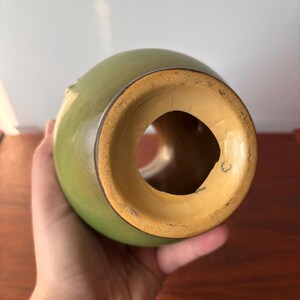 May include: A green ceramic vase with a wide, open base and a rounded body. The interior of the base is a light tan colour, and the rim has a dark brown accent. The vase has a smooth, glossy finish.