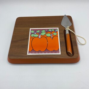 Puede incluir: Una tabla de queso de madera con una baldosa naranja y verde con un diseño de manzanas. La tabla incluye un pequeño cuchillo con mango blanco y una cuerda.