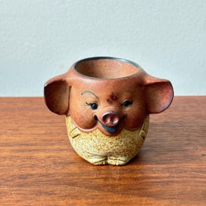 Może przedstawiać: Mały ceramiczny kubek w kształcie świni z dużymi uszami. Górna część jest gładka, czerwonobrązowa, a dolna ma nakrapiane wykończenie w kolorze kremowym i beżowym. Pyszczek świni posiada namalowane czarne oczy, brwi oraz wyraźny ryjek.