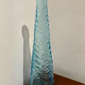 May include: A tall, light blue glass vase with a textured surface that resembles waves. The vase is empty and has a rounded bottom.