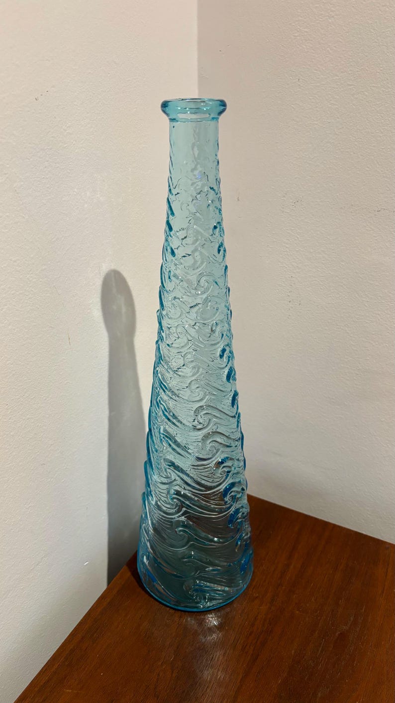 May include: A tall, light blue glass vase with a textured surface that resembles waves. The vase is empty and has a rounded bottom.