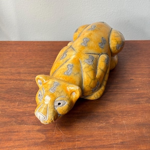 Puede incluir: Una figura de cerámica de un leopardo agachado con un esmalte amarillo mostaza brillante y una textura agrietada. La figura felina presenta manchas de color gris oscuro, abstractas y con forma de reloj de arena en el cuerpo, la cabeza y las extremidades.