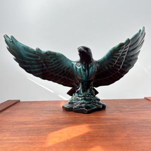 Puede incluir: Una escultura de águila de cerámica con las alas extendidas, acabada en tonos verde azulado y negro. El ave está posada sobre una base texturizada, mostrando patrones de plumas detallados. Es un objeto decorativo.
