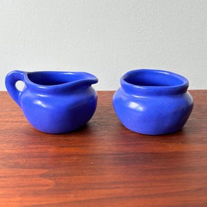 Può includere: Due elementi di servizio in ceramica blu cobalto. Uno è una piccola lattiera con manico e beccuccio. L'altro è una piccola ciotola aperta. Entrambi i pezzi hanno una finitura lucida e poggiano su una superficie in legno scuro.