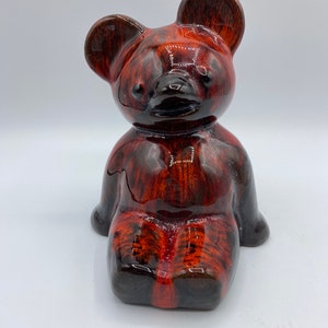May include: A ceramic teddy bear figurine with a brown and orange glaze. The bear is sitting with its legs crossed and has a black nose and eyes.