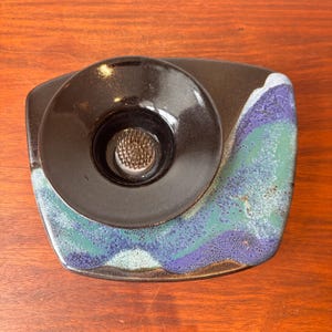 May include: A black ceramic flower frog with a circular design, set on a rectangular base with blue, green, and purple wave-like patterns. The frog has a metal pin holder for arranging flowers.