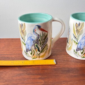 Puede incluir: Dos tazas de cer&aacute;mica blanca con interior turquesa y una ilustraci&oacute;n de garza. La garza es azul con pico rojo, rodeada de follaje verde y amarillo. Las tazas tienen un asa curva y est&aacute;n sobre una superficie de madera.