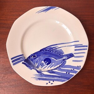 May include: White ceramic plate featuring a blue fish design. The fish is depicted with blue brushstrokes, and the plate has an irregular, slightly square shape. Blue stripes and lines decorate the plate's edge.