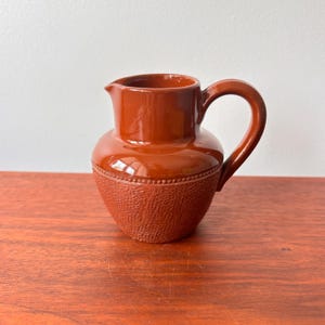 May include: A small, brown ceramic jug with a handle and spout. The jug has a textured lower half and a smooth, glossy upper half. The handle is curved, and the spout is pointed. The jug is sitting on a wooden surface.