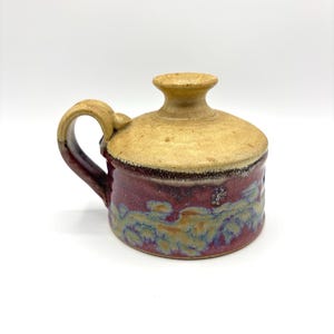 May include: A small, handmade ceramic vase with a brown and red glaze. The vase has a handle and a wide mouth.