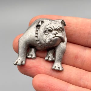 Pode incluir: Estatueta de bulldog prateado com uma superfície texturizada e detalhada. O bulldog está em pé, mostrando suas características distintas. A estatueta é segurada em uma mão contra um fundo cinza.