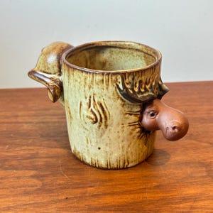 May include: A ceramic mug with a light beige, speckled glaze and a textured finish resembling tree bark. The mug features a three-dimensional sculpted moose head protruding from one side and a sculpted moose tail on the opposite side.
