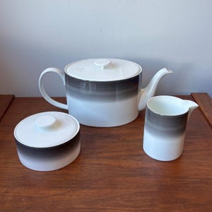 Peut inclure: Un service à thé en céramique blanche composé d'une théière avec couvercle, d'un sucrier avec couvercle et d'un pot à lait. Chaque pièce présente une forme cylindrique lisse avec une bande dégradée gris foncé à noir sur le haut.